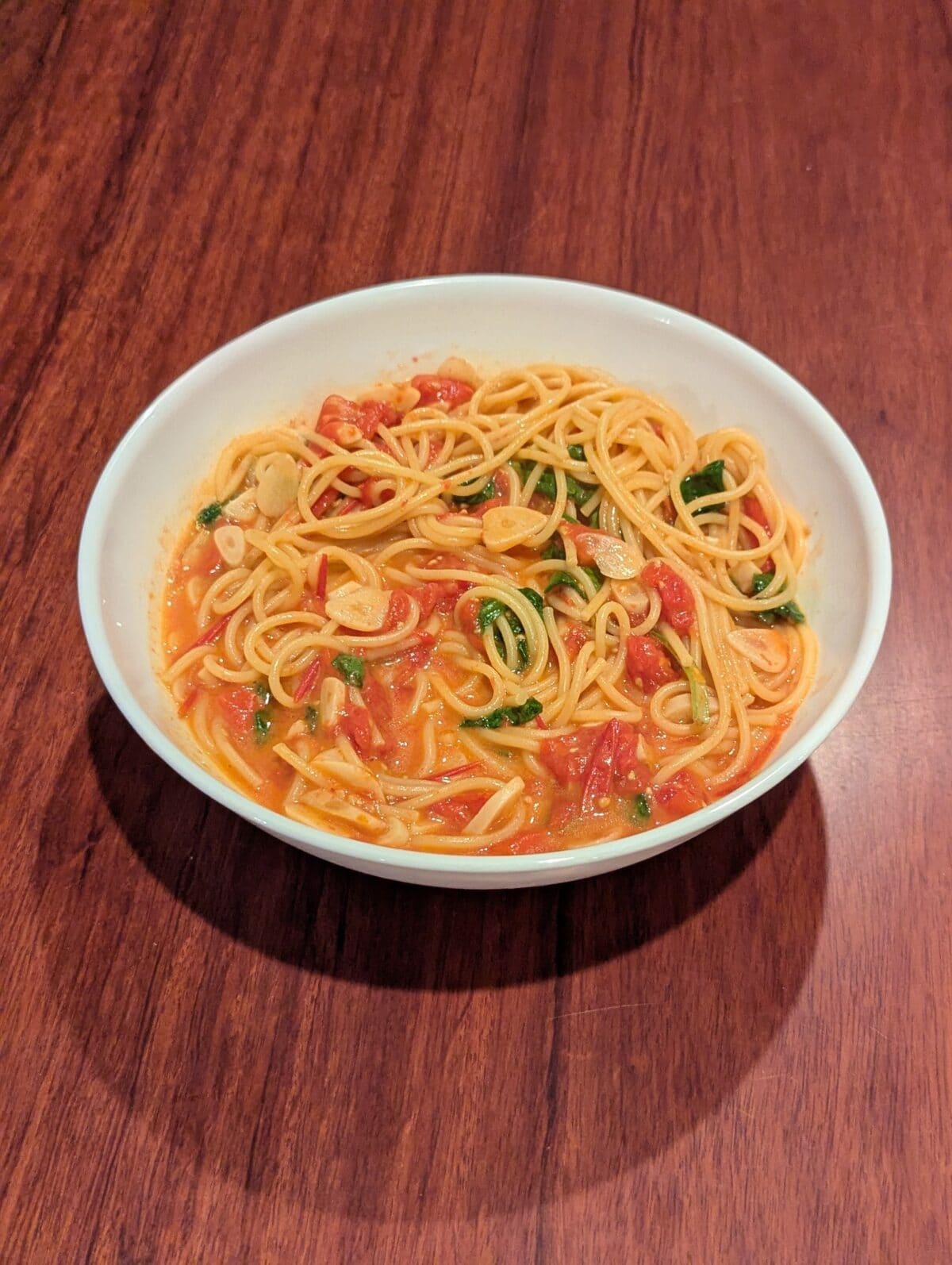 A white bowl filled with spaghetti in a tomato-based sauce with visible slices of garlic and fresh herbs, placed on a wooden table.