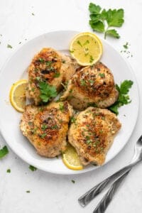 A white plate with four cooked, golden-brown Lemon Yogurt Chicken thighs garnished with chopped herbs and parsley, accompanied by lemon slices. Two forks rest beside the plate on a white surface.