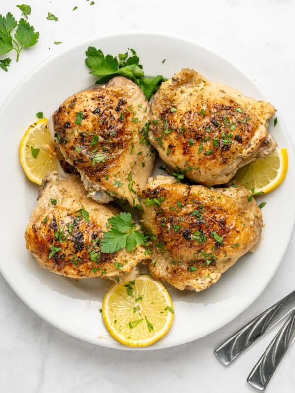Four browned Lemon Yogurt Chicken thighs garnished with chopped parsley, arranged on a white plate with lemon slices and parsley sprigs; two forks sit at the edge of the plate.