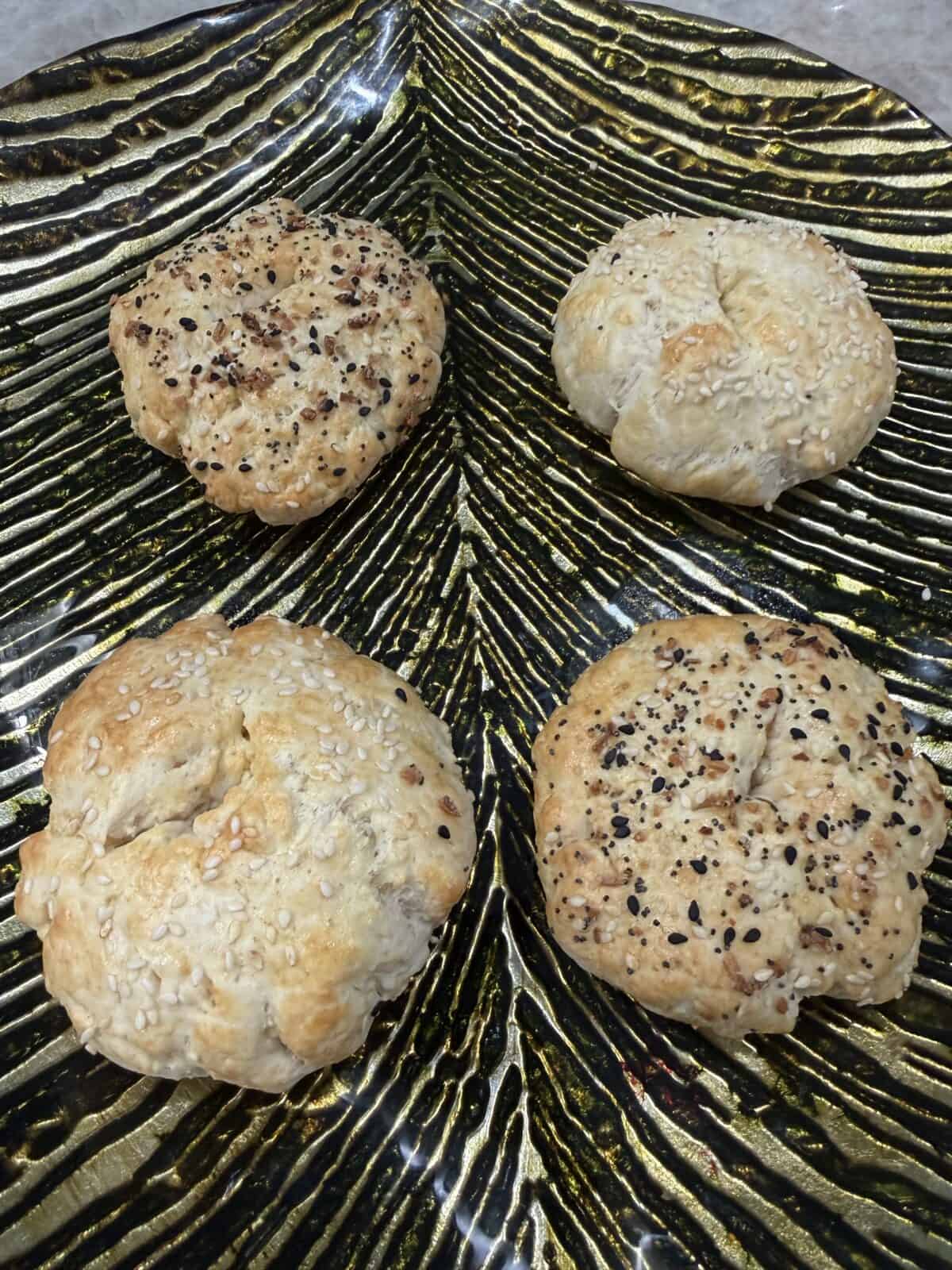Four round, golden-brown Cottage Cheese Bagels topped with sesame seeds and seasoning sit on a metallic, leaf-shaped platter with gold and black stripes.