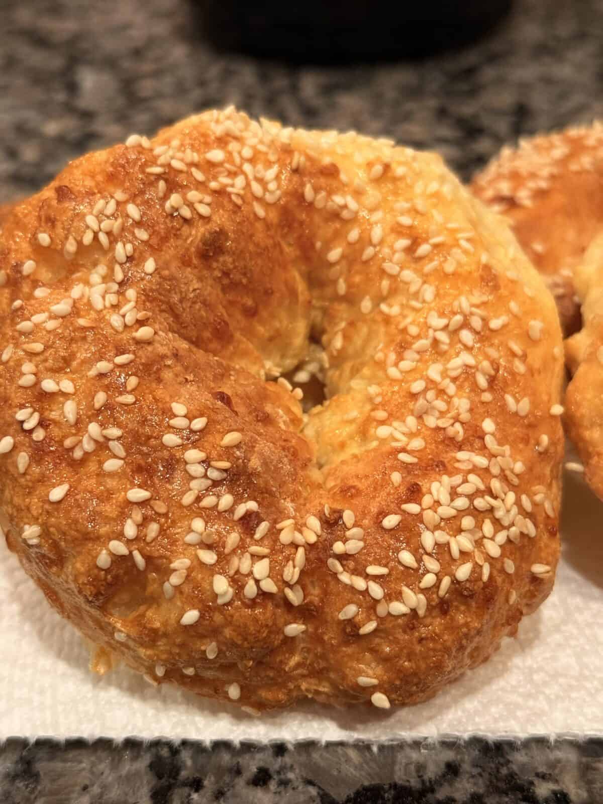 A close-up of a golden brown cottage cheese bagel topped with sesame seeds, resting on a white paper towel on a granite countertop.