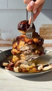 A person uses a fork and spoon to hold a stack of grilled chicken pieces in a pan on a kitchen countertop. In the background, there are kitchen items like a white bowl and a wooden cutting board.