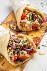 Two gyros filled with grilled meat, fresh tomatoes, red onion slices, crumbled feta, and herbs are served on a wooden board. A creamy sauce is drizzled on top, and the gyros are wrapped in pita bread.