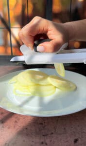 A hand is slicing a white onion using a handheld slicer over a white plate. Sunlight creates soft shadows on the wooden surface, enhancing the kitchen setting's warm ambiance.