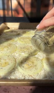 A hand is sprinkling seasoning from a shaker onto sliced onions arranged on top of a layer of rice in a baking dish. The dish is lined with parchment paper and bathed in warm sunlight.