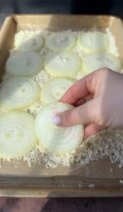 A hand holding a slice of raw onion over a tray filled with multiple onion slices and grated Parmesan cheese. The scene is well-lit, emphasizing the layers and texture of the onions.