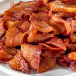 A white plate is filled with crispy, cooked strips of bacon, arranged in a slightly scattered manner. The bacon appears glossy and has a golden-brown color, suggesting it's freshly cooked. The background is a plain, light surface.