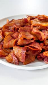 A white plate is filled with crispy, cooked strips of bacon, arranged in a slightly scattered manner. The bacon appears glossy and has a golden-brown color, suggesting it's freshly cooked. The background is a plain, light surface.