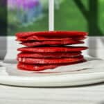 A stack of thin, red fruit leather squares on a white plate with parchment paper. The background shows a blurred view of a window with green trees and a colorful garden outside. Sunlight casts shadows across the table.