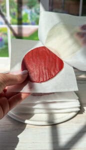 A hand holding a thin, round sheet of red fruit leather on a piece of parchment paper. More layers of fruit leather are stacked below on a plate. Sunlight streams in from the window, illuminating the scene.