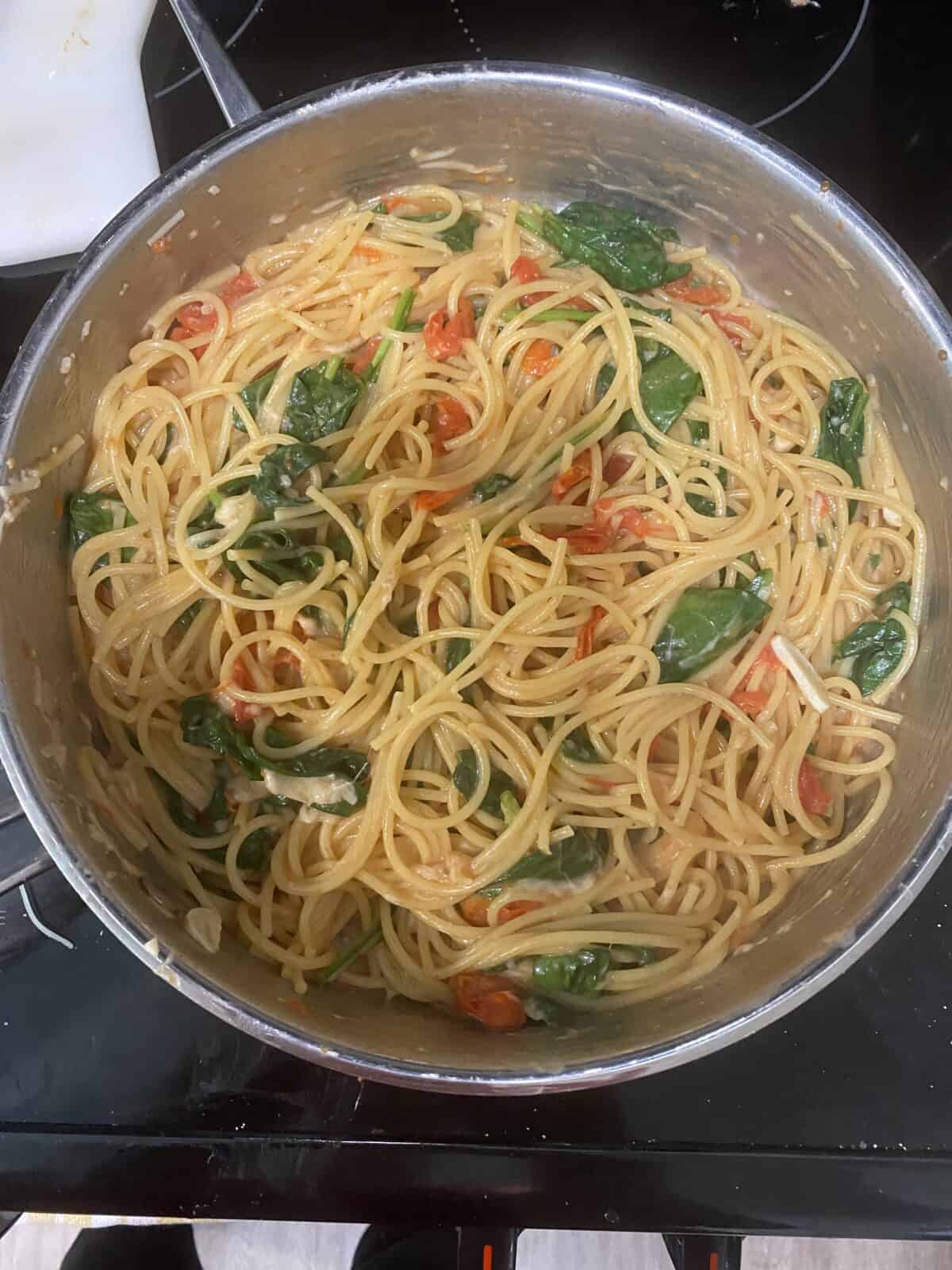 A pot of cooked spaghetti mixed with cherry tomatoes, spinach, and a light sauce, sitting on a stovetop.