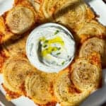 A plate of crispy spiralized onions with golden-brown edges, arranged around a bowl of creamy dip garnished with herbs and olive oil. The onions appear seasoned, and the plate is set against a textured surface.