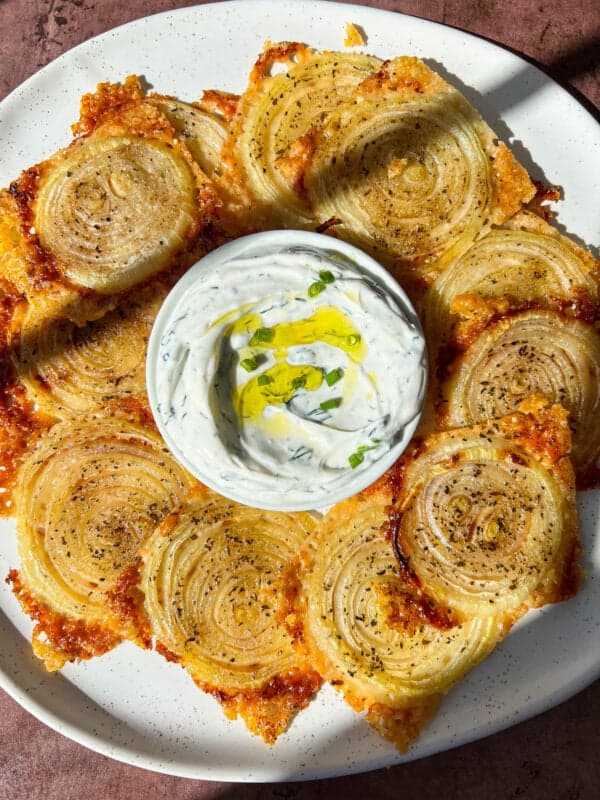 A plate of crispy spiralized onions with golden-brown edges, arranged around a bowl of creamy dip garnished with herbs and olive oil. The onions appear seasoned, and the plate is set against a textured surface.