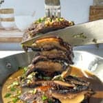 A fork and knife are slicing through a stack of cooked Portobello mushrooms, topped with chopped parsley and garlic, in a pan filled with sauce. The kitchen background is softly blurred.