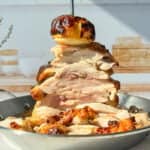 A towering stack of sliced, roasted chicken pieces is skewered on a vertical rod in a pan. A roasted onion tops the stack. The setting includes a bright kitchen with a wooden cutting board and decorative jars in the background.