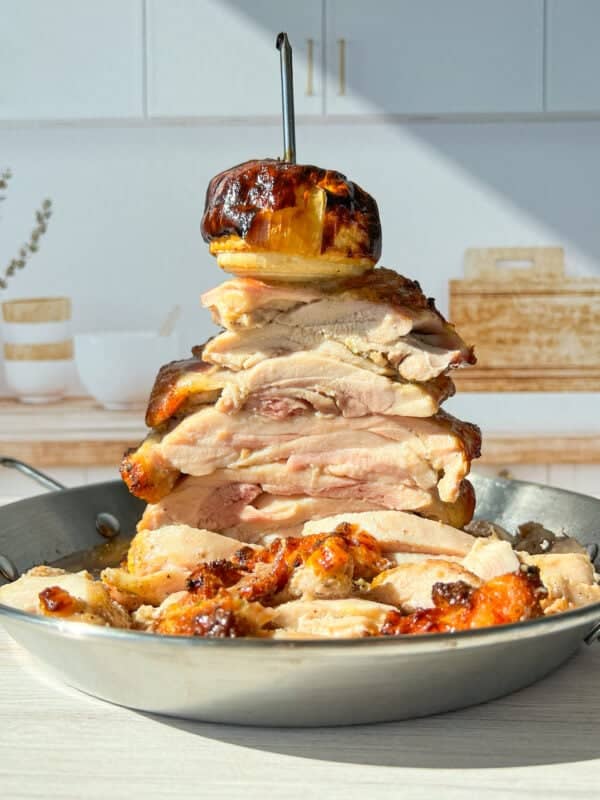 A towering stack of sliced, roasted chicken pieces is skewered on a vertical rod in a pan. A roasted onion tops the stack. The setting includes a bright kitchen with a wooden cutting board and decorative jars in the background.
