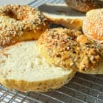 A close-up of several bagels on a cooling rack in bright sunlight. Some are topped with mixed seeds, while one bagel is sliced open, revealing a soft interior. The scene suggests fresh baking and a cozy atmosphere.