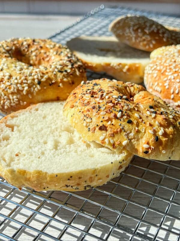 A close-up of several bagels on a cooling rack in bright sunlight. Some are topped with mixed seeds, while one bagel is sliced open, revealing a soft interior. The scene suggests fresh baking and a cozy atmosphere.