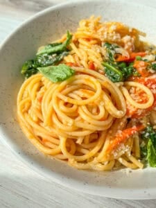 A close-up of a white bowl filled with spaghetti pasta. The pasta is mixed with sautéed spinach, cherry tomatoes, and garnished with grated cheese and a fresh basil leaf. The dish is brightly lit, highlighting the vibrant colors of the ingredients.