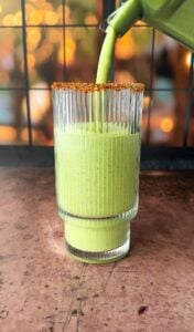 A glass with a chili powder rim is being filled with a green smoothie. The setting appears to be indoors with a window in the background, showcasing a blurred view of trees with warm lighting. The glass stands on a wooden surface.