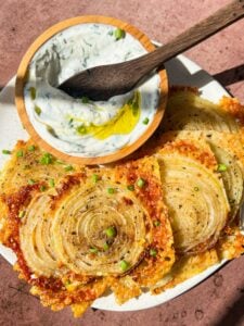 A plate of crispy onion slices, garnished with chopped chives and black pepper, sits next to a small bowl of creamy dip with herbs and a drizzle of olive oil. A wooden spoon rests in the dip. The background is a warm, textured surface.
