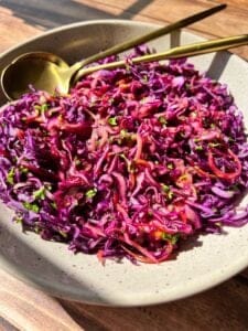 A bowl of vibrant red cabbage slaw with finely shredded purple cabbage, carrots, and herbs, topped with a light vinaigrette. A gold fork and spoon rest beside the dish on a wooden table.