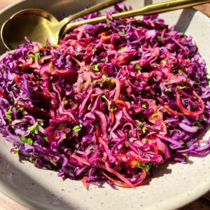 A bowl of vibrant red cabbage slaw with finely shredded purple cabbage, carrots, and herbs, topped with a light vinaigrette. A gold fork and spoon rest beside the dish on a wooden table.