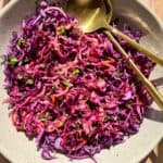 A bowl of colorful red cabbage coleslaw garnished with parsley is served in a light-colored dish. Two gold-colored utensils are placed on the side. The dish is set on a wooden surface, illuminated by natural light.