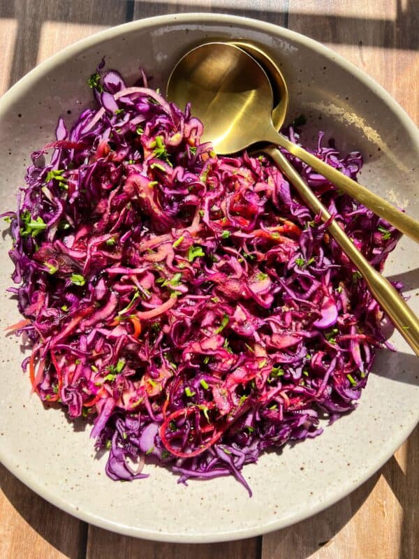 A bowl of colorful red cabbage coleslaw garnished with parsley is served in a light-colored dish. Two gold-colored utensils are placed on the side. The dish is set on a wooden surface, illuminated by natural light.