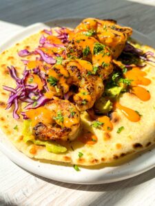 A shrimp taco on a white plate, topped with grilled shrimp, shredded purple cabbage, avocado, fresh herbs, and drizzled with an orange sauce. The taco is on a light wooden table, bathed in bright sunlight.