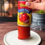 A hand holding an upside-down can of tomato paste, with the paste standing upright on a white plate. A green plant and a black window grid are in the background.