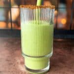 A tall, ridged glass is being filled with a smooth green smoothie. The rim of the glass is coated with a spice mix. The background is softly blurred with warm, golden hues, suggesting evening light through a window.