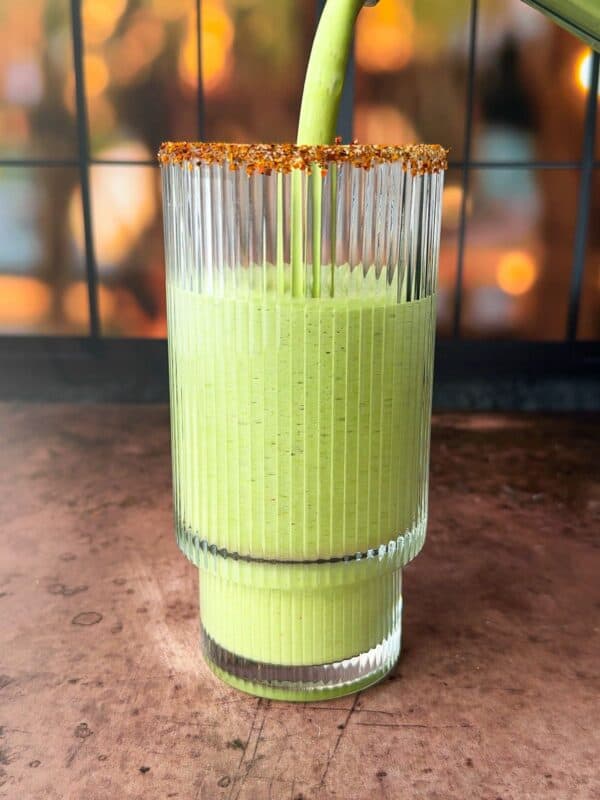 A tall, ridged glass is being filled with a smooth green smoothie. The rim of the glass is coated with a spice mix. The background is softly blurred with warm, golden hues, suggesting evening light through a window.