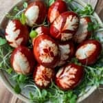 A bowl filled with red-brown dyed eggs featuring intricate leaf patterns, surrounded by fresh green sprouts. The eggs are arranged on a wooden surface, creating a natural, artistic presentation.
