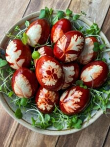 A bowl filled with red-brown dyed eggs featuring intricate leaf patterns, surrounded by fresh green sprouts. The eggs are arranged on a wooden surface, creating a natural, artistic presentation.
