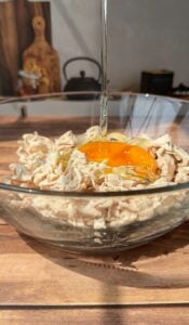 A glass bowl filled with shredded chicken and a raw egg yolk being poured over it, set on a wooden surface with kitchen items blurred in the background.