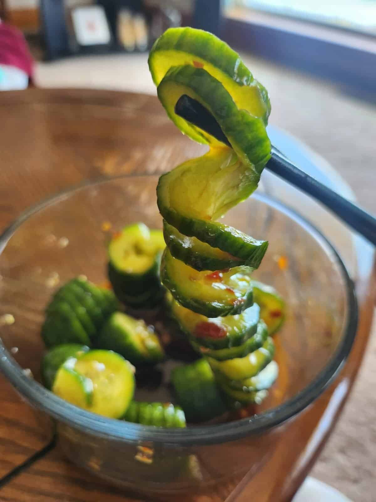 A spiral-cut cucumber is held up by chopsticks over a glass bowl filled with more spiral-cut cucumber slices and light sauce, showcasing a fresh Asian Cucumber Salad set indoors on a wooden surface.