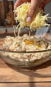 A hand sprinkles shredded cheese into a glass bowl containing shredded chicken and cracked eggs, all set on a wooden surface in bright natural light.