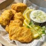 A bowl with golden-brown chicken nuggets, lemon slices, a small cup of creamy dipping sauce, and a sprig of microgreens, served on printed paper in bright natural light.