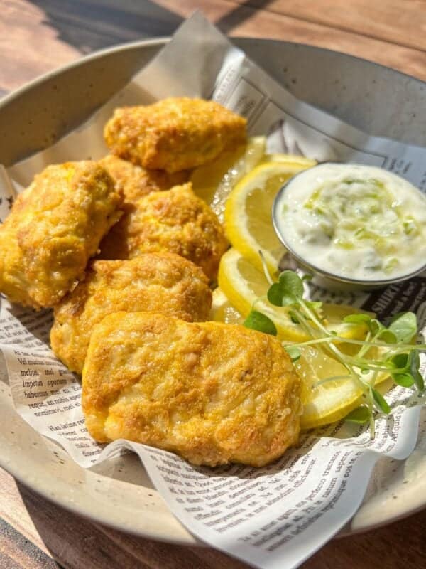 A bowl with golden-brown chicken nuggets, lemon slices, a small cup of creamy dipping sauce, and a sprig of microgreens, served on printed paper in bright natural light.
