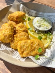 A plate with golden, crispy battered pieces, slices of lemon, microgreens, and a small cup of creamy dipping sauce, all served on newspaper-style paper. Sunlight highlights the texture of the food.
