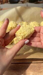 A person holds a rectangular, uncooked snack that appears to be made from instant ramen noodles; more similar snacks rest on a parchment-lined baking sheet in the background.