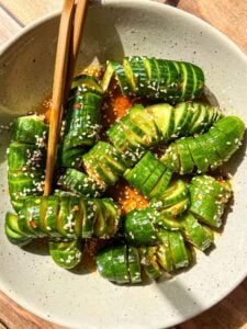 A bowl of spiral-cut cucumbers seasoned with sesame seeds and sliced green onions. The cucumbers are drizzled with a sauce and garnished with sesame seeds. Chopsticks rest on top of the bowl.