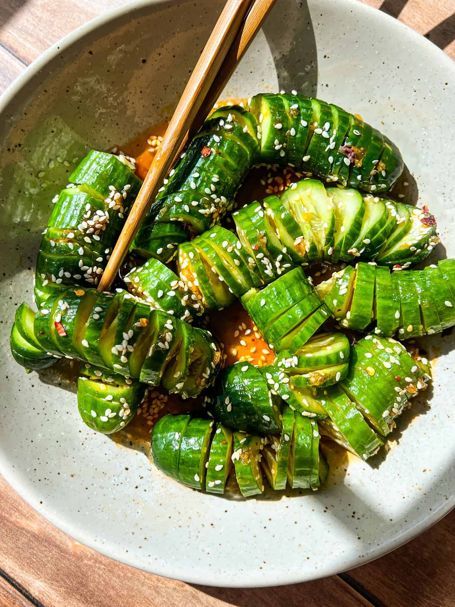 A plate of sliced cucumbers arranged in an S-shape, seasoned with sesame seeds, chili flakes, and a savory sauce. Chopsticks rest on the plate. The dish is bathed in sunlight on a wooden table.