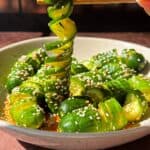 A bowl of spiral-cut cucumbers with a glossy, seasoned dressing, sprinkled with sesame seeds. A hand uses chopsticks to lift a spiral cucumber slice. Background features a softly lit window with grid patterns.
