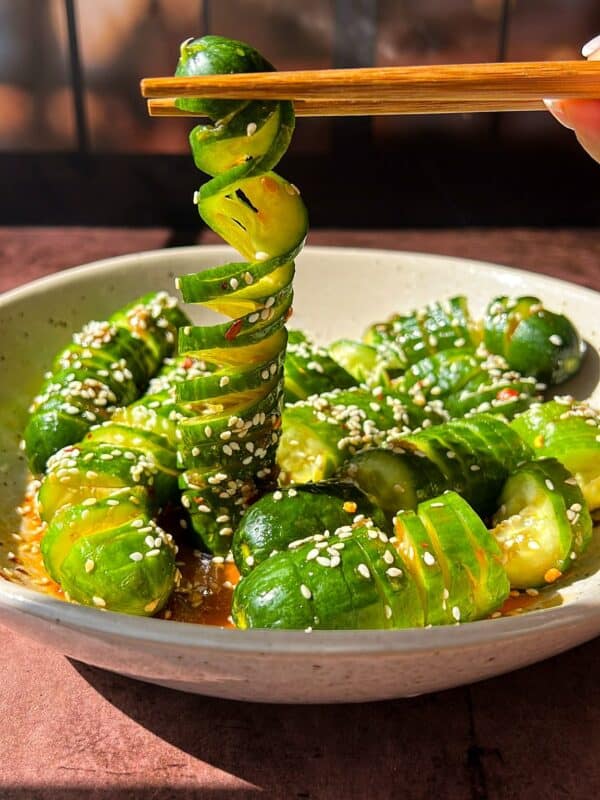 A bowl of spiral-cut cucumbers with a glossy, seasoned dressing, sprinkled with sesame seeds. A hand uses chopsticks to lift a spiral cucumber slice. Background features a softly lit window with grid patterns.
