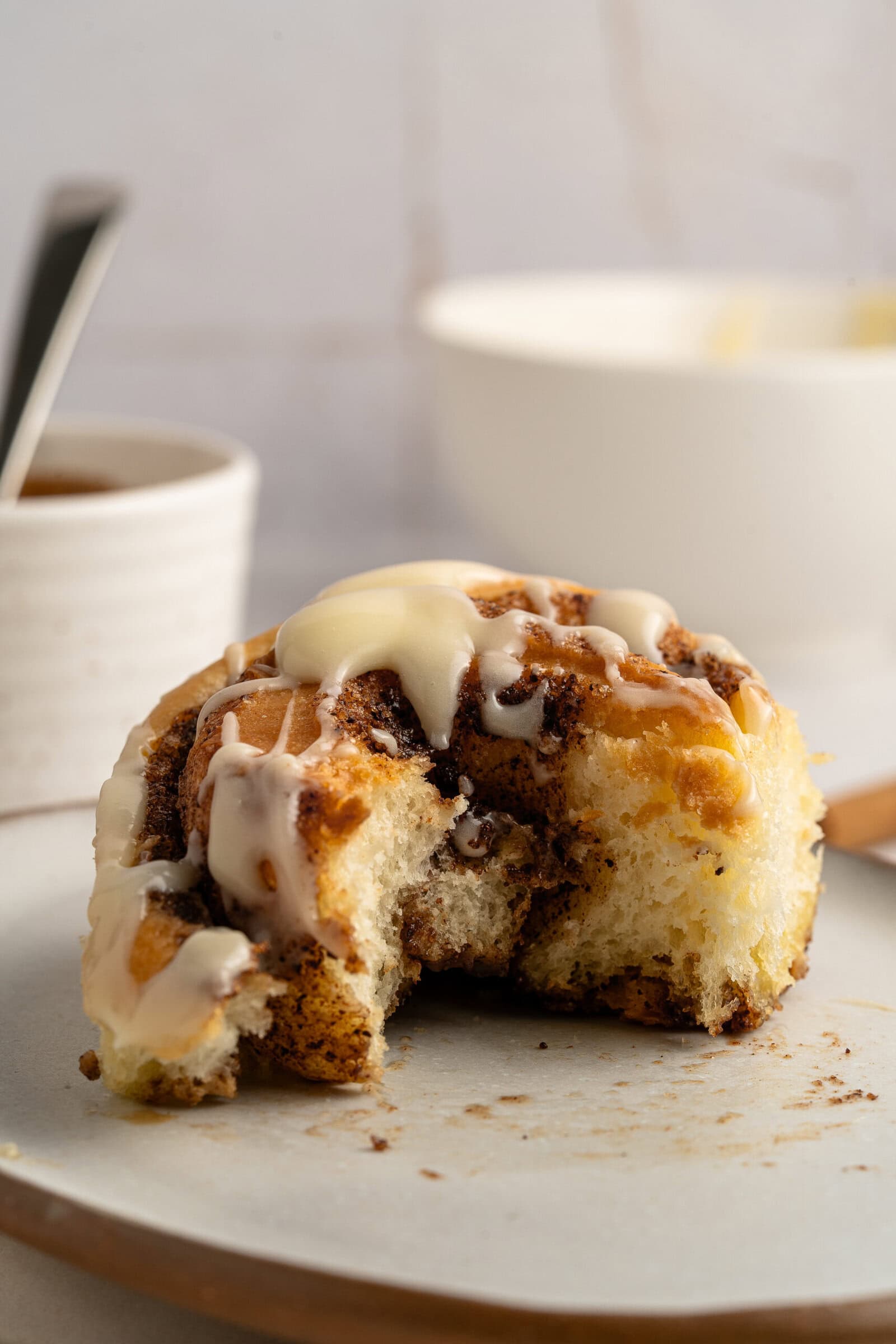 A partially eaten cinnamon roll topped with drizzled icing sits on a plate, with a cup and bowl blurred in the background.