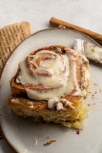 A close-up of a cinnamon roll from a Homemade Cinnamon Rolls Recipe, topped with creamy icing, sits on a gray plate, with a butter knife and an old book page nearby.
