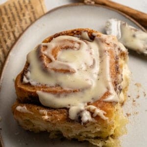 A close-up of a cinnamon roll from a Homemade Cinnamon Rolls Recipe, topped with creamy icing, sits on a gray plate, with a butter knife and an old book page nearby.