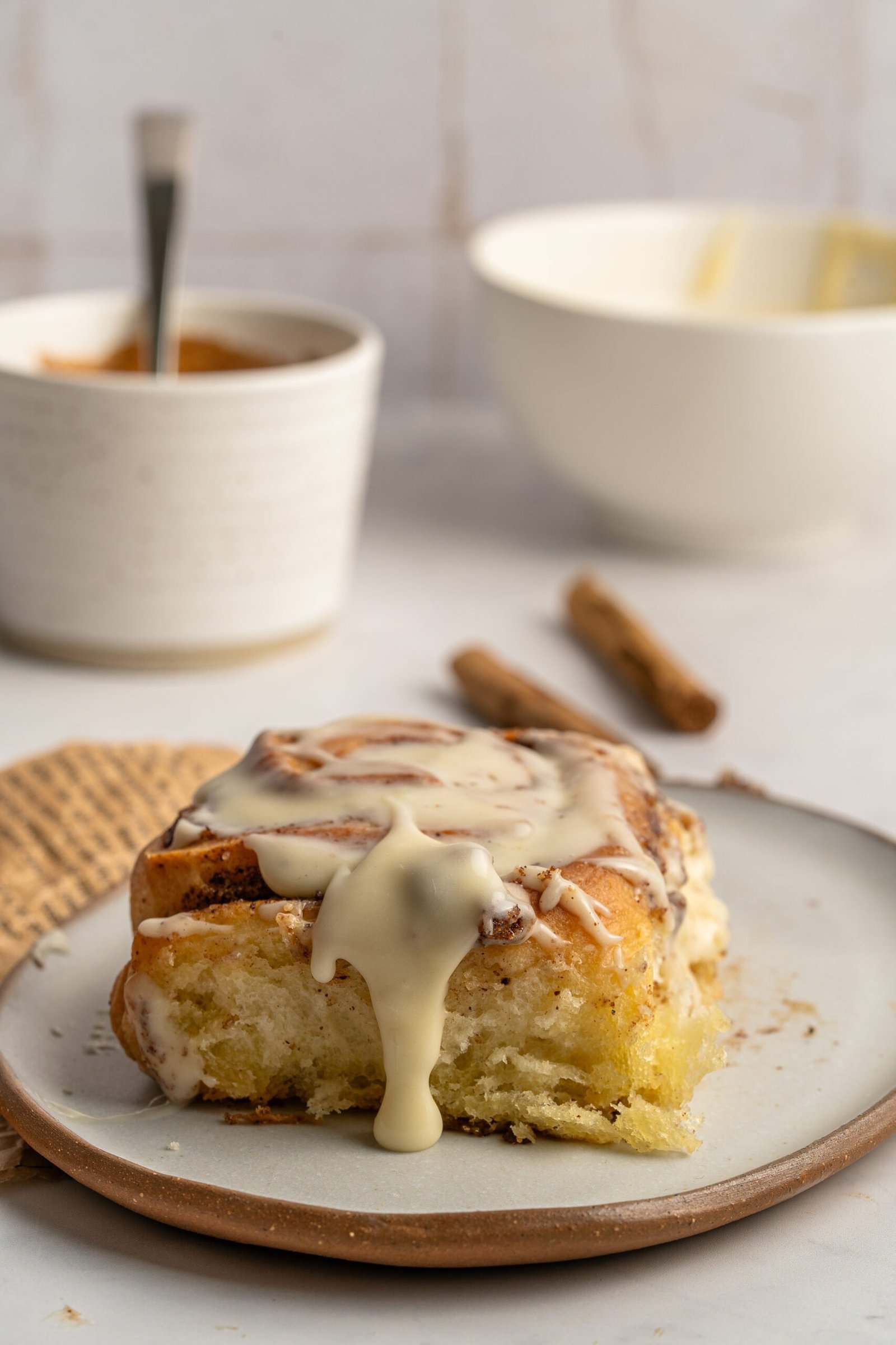 A cinnamon roll with creamy icing dripping down the sides sits on a plate. In the background, there are two bowls and two cinnamon sticks on a light-colored surface.